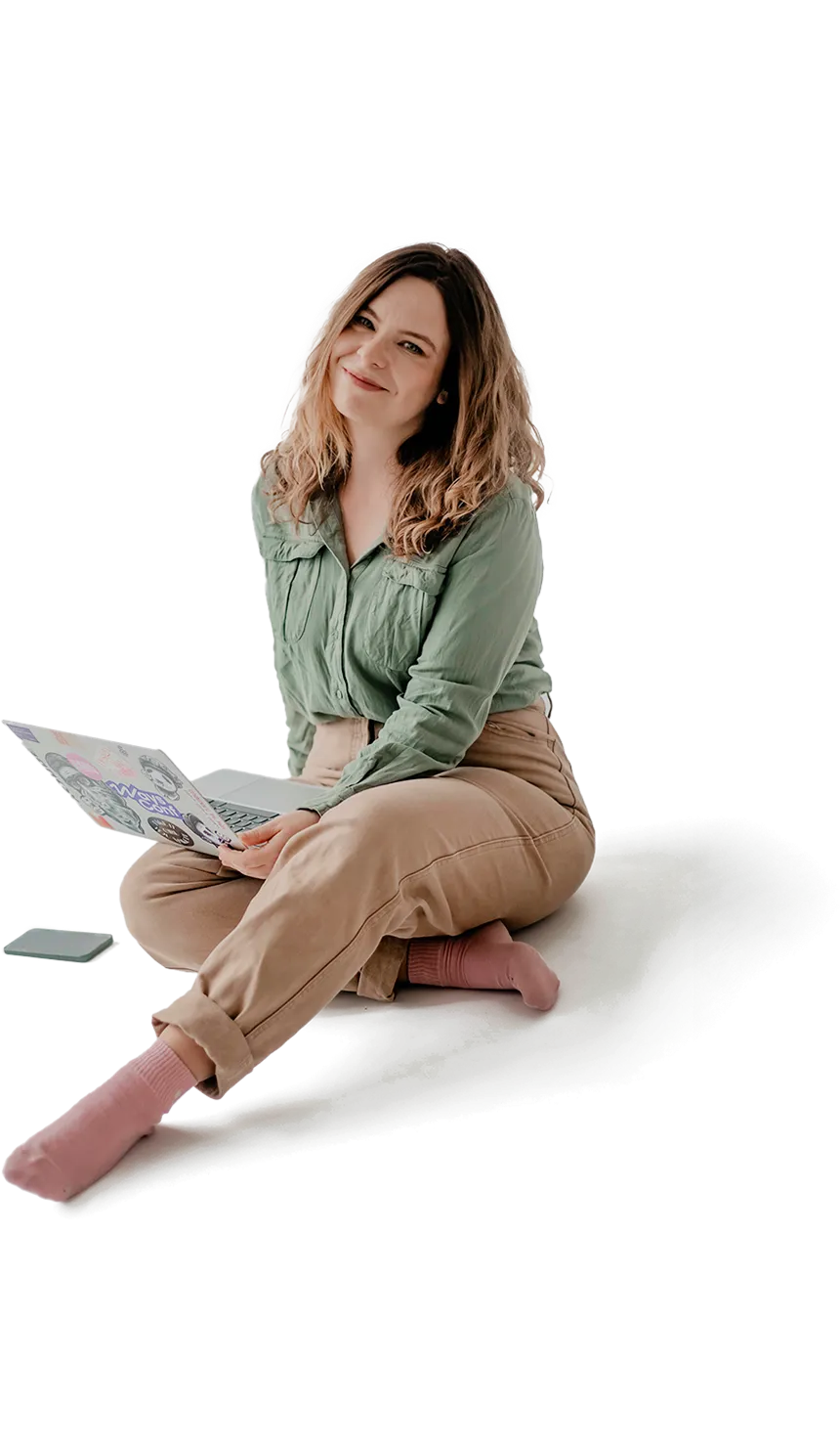 Smiling woman sitting cross-legged on the floor holding a laptop with stickers, wearing a green shirt, beige pants, and pink socks.